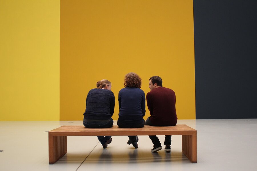Three museum visitors seen from behind, seated on a bench facing a colorful gallery wall